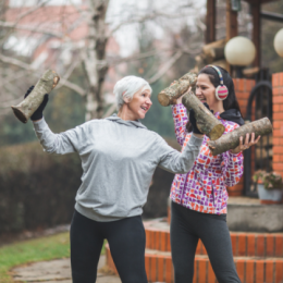 Older woman and younger woman in exercise clothes smiling outside as they lift wood logs