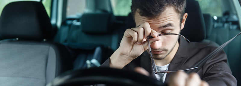 Tired man grabbing his eyes while holding his glasses and clutching steering wheel of car