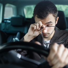 Tired man grabbing his eyes while holding his glasses and clutching steering wheel of car