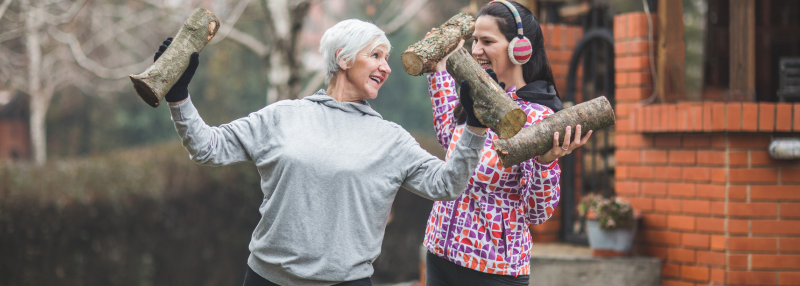 Older woman and younger woman in exercise clothes smiling outside as they lift wood logs