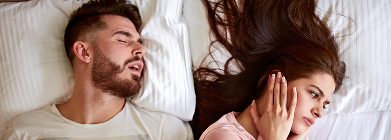 Man sleeping with mouth open presumably snoring with woman next to him with hands over her ears